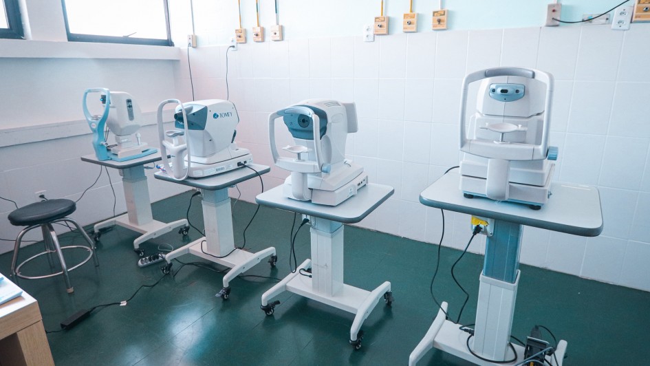 Sala de exames oftalmol&oacute;gicos com quatro equipamentos especializados alinhados lado a lado sobre suportes com rod&iacute;zios. A sala tem piso escuro, paredes claras e pontos de energia organizados na parede.