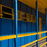 A fotografia mostra uma perspectiva lateral de uma "brizoleta" (escola modular de madeira), localizada no Instituto Professora Gema Angelina Belia, em Porto Alegre.

Elementos Principais
A Estrutura: Trata-se de uma constru&ccedil;&atilde;o alongada feita inteiramente de madeira, com t&aacute;buas horizontais sobrepostas. A arquitetura &eacute; simples e funcional, t&iacute;pica desse modelo educacional hist&oacute;rico.

Cores: A fachada &eacute; pintada em um contraste vibrante de azul e amarelo. As paredes e os pilares de sustenta&ccedil;&atilde;o s&atilde;o azuis, enquanto as molduras das janelas, os corrim&atilde;os da varanda e a parte inferior do telhado s&atilde;o amarelos.

Janelas e Varanda: H&aacute; uma fileira cont&iacute;nua de janelas basculantes de madeira, v&aacute;rias delas abertas para fora. Uma varanda estreita percorre toda a extens&atilde;o da lateral do pr&eacute;dio, protegida por um corrim&atilde;o de madeira amarelo de duas ripas.

Base: A estrutura est&aacute; ligeiramente elevada do ch&atilde;o, apoiada sobre pequenos pilares ou bases de concreto, deixando um v&atilde;o livre entre o piso da escola e o terreno.

Detalhes e Ambiente
Composi&ccedil;&atilde;o: A foto foi tirada em &acirc;ngulo, criando uma linha de fuga que vai da esquerda para a direita, dando profundidade ao corredor da escola.

Entorno: &Agrave; direita, ao fundo, v&ecirc;-se a copa de &aacute;rvores verdes e o c&eacute;u claro, sugerindo um ambiente escolar arborizado. O solo sob a estrutura &eacute; composto por terra e grama rala.

Ilumina&ccedil;&atilde;o: A luz solar incide diretamente sobre a constru&ccedil;&atilde;o, real&ccedil;ando a textura da madeira e as cores vivas da pintura.