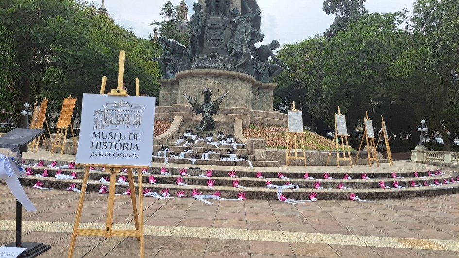 A foto mostra a &aacute;rea em frente a um grande monumento p&uacute;blico, onde est&atilde;o distribu&iacute;das tiras brancas com m&atilde;os cor-de-rosa ao longo dos degraus da estrutura. Ao redor, v&aacute;rios pain&eacute;is e cavaletes exibem informa&ccedil;&otilde;es, e uma placa em destaque identifica o Museu de Hist&oacute;ria J&uacute;lio de Castilhos. 