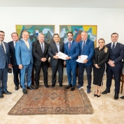 Esta &eacute; uma descri&ccedil;&atilde;o da imagem para fins de acessibilidade:

A fotografia registra um encontro institucional em um gabinete formal, reunindo uma comitiva de lideran&ccedil;as pol&iacute;ticas e representantes setoriais em Bras&iacute;lia.

Elementos Principais
O Centro da A&ccedil;&atilde;o: Ao centro do grupo, o governador Eduardo Leite (vestindo terno azul m&eacute;dio e gravata bord&ocirc;) e o presidente do Senado, Rodrigo Pacheco (vestindo terno cinza escuro), seguram juntos um documento oficial branco. Eles est&atilde;o posicionados no centro da foto, simbolizando a entrega da pauta de reivindica&ccedil;&otilde;es.

A Comitiva: O grupo &eacute; composto por onze pessoas (dez homens e uma mulher), todos vestindo trajes formais (ternos e roupas sociais escuras). Eles est&atilde;o alinhados lado a lado, voltados para a c&acirc;mera com express&otilde;es profissionais e solenes.

Participantes Not&aacute;veis: Al&eacute;m das figuras centrais, &agrave; direita de Leite, um homem segura outro conjunto de documentos. Na extremidade direita, destaca-se uma mulher vestindo um conjunto preto de saia e jaqueta.

Cen&aacute;rio e Ambiente
Decora&ccedil;&atilde;o: O ambiente &eacute; elegante e s&oacute;brio. Atr&aacute;s do grupo, h&aacute; dois quadros grandes de arte abstrata com tons de verde, azul e terra, emoldurados em madeira. O piso &eacute; de pedra clara, com um tapete persa retangular de cores quentes (vermelho e azul) posicionado sob os p&eacute;s das autoridades centrais.

Ilumina&ccedil;&atilde;o: A luz &eacute; uniforme e clara, vinda de fontes embutidas no teto, sem sombras marcadas, caracter&iacute;stica de gabinetes oficiais.

Contexto
A imagem ilustra a articula&ccedil;&atilde;o pol&iacute;tica do governo ga&uacute;cho junto ao Legislativo Federal, buscando o avan&ccedil;o de projetos de lei para a renegocia&ccedil;&atilde;o de d&iacute;vidas do setor agropecu&aacute;rio do Rio Grande do Sul.