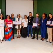 A fotografia registra um grupo de 13 pessoas perfiladas lado a lado em um ambiente interno, celebrando a entrega de certificados do Selo Empresa Parceira da Diversidade Sexual e de G&ecirc;nero.

Elementos Principais
Os Integrantes: O grupo &eacute; diversificado em g&ecirc;nero, idade e etnia. A maioria segura um certificado horizontal com bordas coloridas (nas cores do arco-&iacute;ris), exibindo-os para a c&acirc;mera.

Vestimentas: As roupas variam entre o estilo social e o casual. Destacam-se uma mulher &agrave; esquerda com uma saia longa em degrad&ecirc; colorido, um homem com cabelos descoloridos, e diversas pessoas em trajes executivos (blazers e camisas sociais).

Posicionamento: Eles formam uma linha semicircular em um palco ou espa&ccedil;o de eventos com piso claro. Ao fundo, h&aacute; uma parede verde escura &agrave; direita e uma tela de proje&ccedil;&atilde;o branca retr&aacute;til ao centro, que est&aacute; vazia.

Detalhes e Ambiente
Sinaliza&ccedil;&atilde;o: &Agrave; esquerda, h&aacute; um banner branco (parcialmente vis&iacute;vel) com o bras&atilde;o do Estado do Rio Grande do Sul.

Composi&ccedil;&atilde;o: No canto inferior direito, v&ecirc;-se parte das m&atilde;os e do celular de uma pessoa que tamb&eacute;m fotografa o momento, conferindo uma sensa&ccedil;&atilde;o de evento movimentado.

Ilumina&ccedil;&atilde;o: A luz &eacute; frontal e clara, t&iacute;pica de audit&oacute;rios, garantindo visibilidade a todos os rostos e aos documentos entregues.