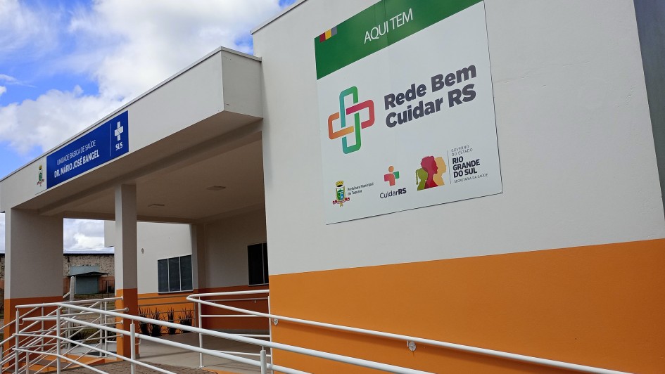 Entrada de unidade de sa&uacute;de com arquitetura em branco e laranja, acompanhada de rampa acess&iacute;vel. Um painel destaca o programa Rede Bem Cuidar RS com logotipos institucionais. O pr&eacute;dio &eacute; t&eacute;rreo e possui estrutura simples e clara.