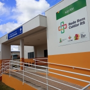 Entrada de unidade de sa&uacute;de com arquitetura em branco e laranja, acompanhada de rampa acess&iacute;vel. Um painel destaca o programa Rede Bem Cuidar RS com logotipos institucionais. O pr&eacute;dio &eacute; t&eacute;rreo e possui estrutura simples e clara.