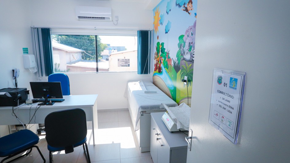 Consult&oacute;rio infantil com decora&ccedil;&atilde;o tem&aacute;tica colorida em uma das paredes, incluindo ilustra&ccedil;&otilde;es de animais e natureza. O espa&ccedil;o possui maca, mesa com computador, cadeiras e janela ampla com cortinas curtas, al&eacute;m de ilumina&ccedil;&atilde;o branca no teto.