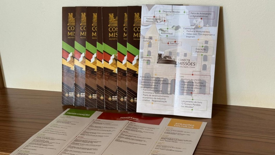 Sobre uma prateleira de madeira, apoiada junto a uma parede clara, est&atilde;o dispostos materiais impressos do projeto &ldquo;Conecta Miss&otilde;es&rdquo;. Na parte de tr&aacute;s, v&aacute;rios folhetos verticais aparecem alinhados lado a lado, com fundo escuro, o t&iacute;tulo &ldquo;Conecta Miss&otilde;es&rdquo; em destaque na parte superior e uma imagem de arquitetura hist&oacute;rica na parte inferior, acompanhada por elementos gr&aacute;ficos em verde e vermelho. &Agrave; frente deles, um material aberto ocupa toda a largura da prateleira, exibindo textos organizados em colunas com cabe&ccedil;alhos coloridos e, na parte inferior, uma fotografia de um conjunto arquitet&ocirc;nico hist&oacute;rico. Ao lado, um painel dobr&aacute;vel apresenta um mapa ilustrado com nomes de localidades, atra&ccedil;&otilde;es e atividades, combinando textos, &iacute;cones e imagens, compondo uma apresenta&ccedil;&atilde;o visual informativa e organizada sobre o projeto.

