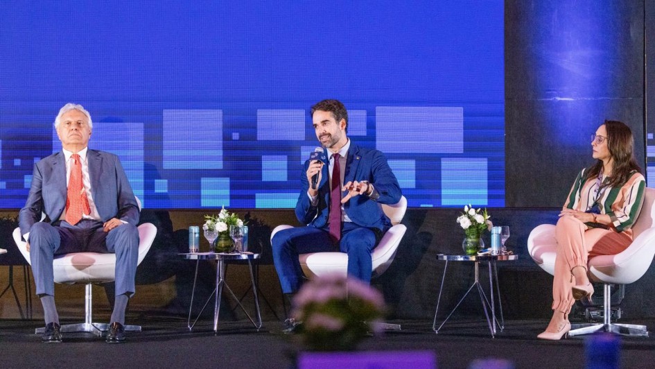 Tr&ecirc;s pessoas est&atilde;o sentadas em poltronas no palco de um evento. Um homem de terno cinza e gravata vermelha est&aacute; &agrave; esquerda, outro homem de terno azul fala ao microfone no centro, e uma mulher com roupa colorida est&aacute; &agrave; direita. Atr&aacute;s deles h&aacute; um grande painel azul com textos.