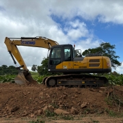 A imagem mostra uma escavadeira hidr&aacute;ulica amarela posicionada sobre um terreno de terra solta. A m&aacute;quina est&aacute; com a concha voltada para baixo, em contato com o solo, e possui esteiras met&aacute;licas apoiadas sobre o amontoado de terra.