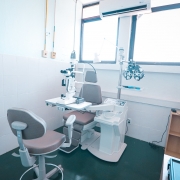 A imagem exibe um consult&oacute;rio oftalmol&oacute;gico equipado com cadeira de exame, l&acirc;mpada de fenda, refrator e mesa ajust&aacute;vel. H&aacute; duas janelas amplas que iluminam a sala, al&eacute;m de ar-condicionado instalado acima delas. 