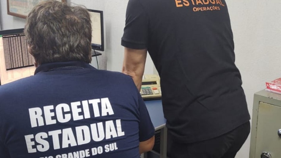 Dois homens de costas, um sentado e outro em p&eacute;, vestindo camisas com a inscri&atilde;o Receita Estadual Rio Grande do Sul e Opera&ccedil;&otilde;es, analisam computadores