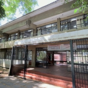 Foto mostra fachada da Escola Uruguai, pr&eacute;dio de dois andares em linha reta. Na frente h&aacute; arvores e um espa&ccedil;o com sombra. 