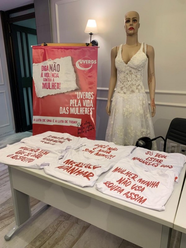 A fotografia apresenta uma instalação de impacto visual para uma campanha de conscientização contra a violência doméstica, organizada pela UVERGS (União dos Vereadores do Rio Grande do Sul).

Elementos Principais
O Manequim: À direita, há um manequim feminino realista vestindo um vestido de noiva branco rendado. O manequim apresenta marcas que simulam violência física: hematomas roxos ao redor dos olhos e "sangue" escorrendo pelo canto da boca, pescoço e braços.

O Banner: À esquerda do manequim, um banner vertical vermelho e branco traz as frases: "DIGA NÃO À VIOLÊNCIA CONTRA A MULHER!" e "UVERGS PELA VIDA DAS MULHERES". No rodapé do banner, lê-se: "A LUTA DE UMA É A LUTA DE TODAS!".

As Camisetas: Em primeiro plano, sobre uma mesa branca, estão dispostas várias camisetas brancas dobradas. Cada uma exibe, em letras vermelhas, frases que reproduzem abusos psicológicos e ameaças comuns em contextos de violência doméstica, como:

"Merece apanhar"

"Mulher minha não usa roupa assim"

"Você não é nada sem mim!!!"

"Só pode estar naqueles dias"

Detalhes e Ambiente
Cenário: A instalação está montada em um ambiente interno com paredes claras e molduras clássicas (boiseries). Ao fundo, vê-se uma porta de madeira escura e uma arandela de parede acesa, que ilumina o manequim de cima para baixo.

Composição: A imagem é organizada para que o olhar do espectador percorra primeiro as frases violentas na mesa, suba para o banner de denúncia e termine no impacto visual do manequim "ferido".

Propósito: A cena utiliza o contraste entre o vestido de noiva (símbolo de união e celebração) e os sinais de agressão para chocar e gerar reflexão sobre a violência oculta nas relações.