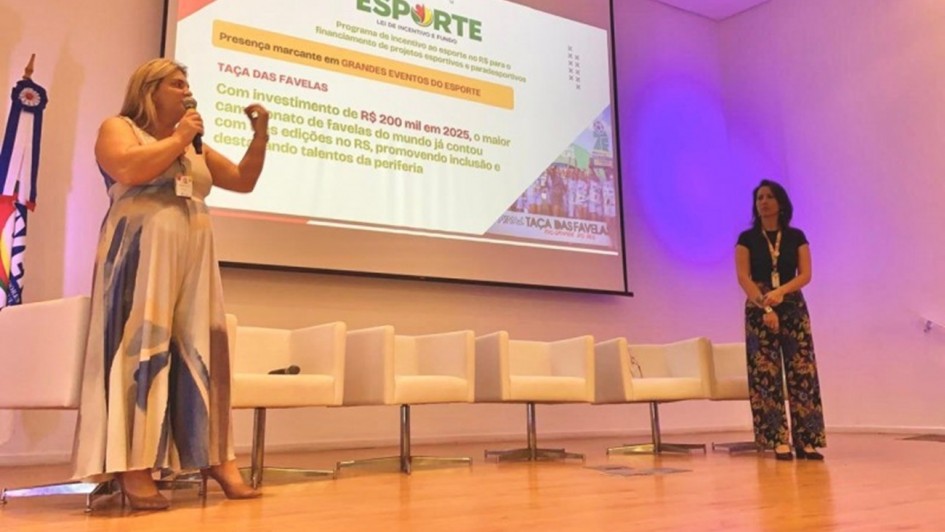 A fotografia registra uma apresenta&ccedil;&atilde;o institucional realizada em um palco de audit&oacute;rio, focada em projetos de incentivo ao esporte.

Elementos Principais
A Oradora (Nathalia, &agrave; esquerda): Uma mulher loira, vestindo um vestido longo com estampas abstratas em tons de azul e bege, fala ao microfone. Ela est&aacute; de p&eacute; no lado esquerdo do palco, gesticulando com a m&atilde;o esquerda enquanto apresenta.

A Assistente/Apoio (Carla, &agrave; direita): Uma mulher de cabelos escuros, vestindo uma blusa preta e cal&ccedil;a com estampa floral colorida, est&aacute; de p&eacute; no lado direito do palco, observando a apresenta&ccedil;&atilde;o. Ela utiliza um crach&aacute; institucional.

A Proje&ccedil;&atilde;o: Ao fundo, uma grande tela exibe um slide do programa "Pr&oacute;-Esporte" (Lei de Incentivo e Fundo). O texto em destaque menciona a "TA&Ccedil;A DAS FAVELAS" e cita um investimento de R$ 200 mil em 2025, descrevendo-o como o maior campeonato de favelas do mundo para promover inclus&atilde;o e talentos da periferia.

Detalhes e Ambiente
Cen&aacute;rio: O palco possui piso de madeira clara e brilhante. Atr&aacute;s das oradoras, h&aacute; uma fileira de cinco poltronas brancas modernas e quadradas, que est&atilde;o vazias. No canto esquerdo, &eacute; vis&iacute;vel parte de uma bandeira oficial em um mastro.

Ilumina&ccedil;&atilde;o: O audit&oacute;rio tem paredes brancas e uma ilumina&ccedil;&atilde;o c&ecirc;nica que projeta um feixe de luz circular arroxeada na parede ao fundo, &agrave; direita da tela.

Composi&ccedil;&atilde;o: A foto foi tirada de um &acirc;ngulo baixo (de baixo para cima), o que d&aacute; destaque e amplitude ao palco e &agrave; tela de proje&ccedil;&atilde;o.