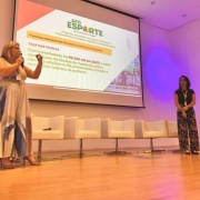 A fotografia registra uma apresenta&ccedil;&atilde;o institucional realizada em um palco de audit&oacute;rio, focada em projetos de incentivo ao esporte.

Elementos Principais
A Oradora (Nathalia, &agrave; esquerda): Uma mulher loira, vestindo um vestido longo com estampas abstratas em tons de azul e bege, fala ao microfone. Ela est&aacute; de p&eacute; no lado esquerdo do palco, gesticulando com a m&atilde;o esquerda enquanto apresenta.

A Assistente/Apoio (Carla, &agrave; direita): Uma mulher de cabelos escuros, vestindo uma blusa preta e cal&ccedil;a com estampa floral colorida, est&aacute; de p&eacute; no lado direito do palco, observando a apresenta&ccedil;&atilde;o. Ela utiliza um crach&aacute; institucional.

A Proje&ccedil;&atilde;o: Ao fundo, uma grande tela exibe um slide do programa "Pr&oacute;-Esporte" (Lei de Incentivo e Fundo). O texto em destaque menciona a "TA&Ccedil;A DAS FAVELAS" e cita um investimento de R$ 200 mil em 2025, descrevendo-o como o maior campeonato de favelas do mundo para promover inclus&atilde;o e talentos da periferia.

Detalhes e Ambiente
Cen&aacute;rio: O palco possui piso de madeira clara e brilhante. Atr&aacute;s das oradoras, h&aacute; uma fileira de cinco poltronas brancas modernas e quadradas, que est&atilde;o vazias. No canto esquerdo, &eacute; vis&iacute;vel parte de uma bandeira oficial em um mastro.

Ilumina&ccedil;&atilde;o: O audit&oacute;rio tem paredes brancas e uma ilumina&ccedil;&atilde;o c&ecirc;nica que projeta um feixe de luz circular arroxeada na parede ao fundo, &agrave; direita da tela.

Composi&ccedil;&atilde;o: A foto foi tirada de um &acirc;ngulo baixo (de baixo para cima), o que d&aacute; destaque e amplitude ao palco e &agrave; tela de proje&ccedil;&atilde;o.