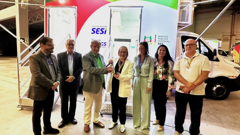 Um grupo de pessoas est&aacute; alinhado em frente a uma unidade m&oacute;vel de sa&uacute;de. Todas est&atilde;o posicionadas sob o toldo lateral e diante das escadas de acesso. A identidade visual do SESI, FIERGS e governo estadual aparece ao fundo, destacada pela ilumina&ccedil;&atilde;o da estrutura.
