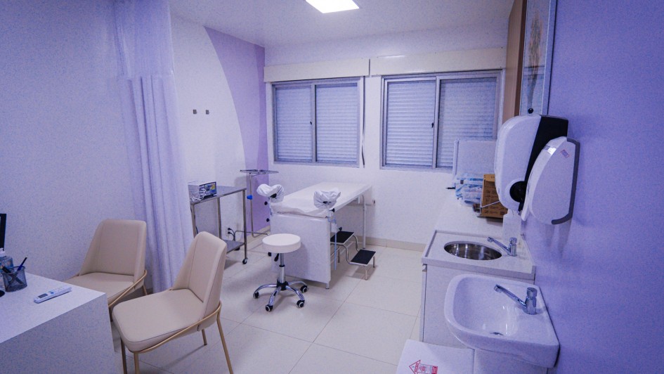 Foto mostra ambulat&oacute;rio com mesas para atendimento, maca e pias para lavaem das m&atilde;os. As paredes e m&oacute;veis s&atilde;o na tonalidade lil&aacute;s.