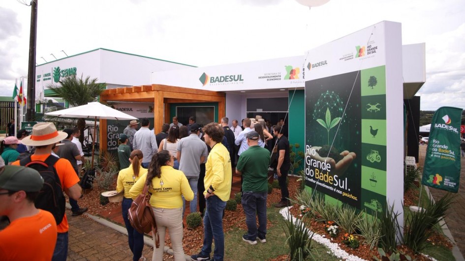 A imagem mostra o estande do Badesul em um evento ao ar livre, identificado por pain&eacute;is grandes com a marca da institui&ccedil;&atilde;o e elementos gr&aacute;ficos relacionados ao desenvolvimento econ&ocirc;mico e sustent&aacute;vel. Um grupo de visitantes circula e conversa na &aacute;rea externa, onde tamb&eacute;m h&aacute; bandeiras, estandartes e um bal&atilde;o infl&aacute;vel com o logotipo do Badesul. 