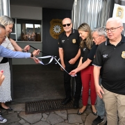 Esta &eacute; uma descri&ccedil;&atilde;o da imagem para fins de acessibilidade:

A fotografia registra o ato inaugural de duas novas delegacias especializadas no munic&iacute;pio de Rio Grande, com autoridades e agentes da Pol&iacute;cia Civil participando do desatamento da fita.

Elementos Principais
O Ato: Sete pessoas est&atilde;o posicionadas na entrada de um pr&eacute;dio. Tr&ecirc;s pessoas &agrave; esquerda e duas &agrave; direita seguram e puxam uma fita inaugural de tecido branco e preto, simbolizando a abertura oficial das unidades.

Os Participantes:

&Agrave; esquerda: Um homem de camisa azul clara e uma mulher de blusa branca rendada puxam a fita com entusiasmo.

Ao centro: Um homem de &oacute;culos escuros, vestindo a camiseta polo preta oficial da Pol&iacute;cia Civil, observa o ato com um sorriso.

&Agrave; direita: Uma mulher de cal&ccedil;a vermelha e camiseta polo da Pol&iacute;cia Civil ajuda a conduzir a fita, acompanhada por outros dois homens tamb&eacute;m uniformizados.

Identifica&ccedil;&atilde;o: Na parede ao fundo, v&ecirc;-se o distintivo dourado da Pol&iacute;cia Civil do RS. &Agrave; direita, h&aacute; um painel (backdrop) com o padr&atilde;o "checkboard" (xadrez) em preto e branco, alternando o bras&atilde;o da institui&ccedil;&atilde;o e as palavras "POL&Iacute;CIA CIVIL".

Cen&aacute;rio e Ambiente
Local: A entrada possui piso de pedras irregulares e uma grade met&aacute;lica de prote&ccedil;&atilde;o. Ao lado da porta, h&aacute; um vaso com uma planta ornamental verde.

Atmosfera: O clima &eacute; de celebra&ccedil;&atilde;o e dever cumprido. Algumas pessoas ao lado esquerdo aparecem aplaudindo o momento do desenlace da fita.

Ilumina&ccedil;&atilde;o: A luz &eacute; natural e intensa (dia claro), criando sombras suaves no ch&atilde;o e destacando as texturas das roupas e da fachada.