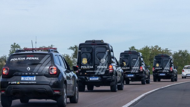 Esta &eacute; uma descri&ccedil;&atilde;o da imagem para fins de acessibilidade:

A fotografia registra um comboio de viaturas da Pol&iacute;cia Penal do Rio Grande do Sul em deslocamento por uma rodovia, ilustrando a marca de mais de 80 mil escoltas realizadas em 2025.

Elementos Principais
O Comboio: Quatro viaturas oficiais pretas est&atilde;o alinhadas em uma estrada de pista simples. As viaturas seguem em fila indiana, afastando-se da c&acirc;mera (vis&atilde;o traseira).

Tipos de Ve&iacute;culos:

Em primeiro plano, &agrave; esquerda, h&aacute; um utilit&aacute;rio modelo Renault Duster com o giroflex vermelho e azul ligado. Na parte superior do vidro traseiro, l&ecirc;-se "POL&Iacute;CIA PENAL" e, abaixo da placa, a identifica&ccedil;&atilde;o "1&ordm; DPR".

Logo &agrave; frente, seguem tr&ecirc;s furg&otilde;es de maior porte (estilo viaturas de transporte de presos ou escolta t&aacute;tica), tamb&eacute;m pretos, com grades nas janelas laterais e traseiras e a inscri&ccedil;&atilde;o "POL&Iacute;CIA PENAL" em destaque na lataria traseira.

Identifica&ccedil;&atilde;o: Todas as viaturas possuem o bras&atilde;o dourado da Pol&iacute;cia Penal e a orienta&ccedil;&atilde;o de seguran&ccedil;a "MANTENHA DIST&Acirc;NCIA" escrita em letras brancas.

Cen&aacute;rio e Ambiente
Rodovia: A estrada possui asfalto escuro com sinaliza&ccedil;&atilde;o de faixa branca cont&iacute;nua &agrave; direita. O comboio ocupa a faixa principal, enquanto ao fundo, &agrave; direita, um carro de passeio branco trafega no sentido oposto.

Paisagem: As margens da rodovia s&atilde;o compostas por vegeta&ccedil;&atilde;o rasteira e &aacute;rvores verdes sob um c&eacute;u claro e levemente nublado.

Composi&ccedil;&atilde;o: A foto utiliza uma perspectiva de profundidade, focando no alinhamento das viaturas, o que transmite uma sensa&ccedil;&atilde;o de organiza&ccedil;&atilde;o, seguran&ccedil;a e prontid&atilde;o operacional.