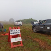 Imagem mostra uma estrada de terra e uma placa laranja onde est&aacute; escrito Fiscaliza&ccedil;&atilde;o Agropecu&aacute;ria. H&aacute; caminhonetes paradas atr&aacute;s e um t&eacute;cnico fazendo a fiscaliza&ccedil;&atilde;o. O dia est&aacute; com cerra&ccedil;&atilde;o baixa. 