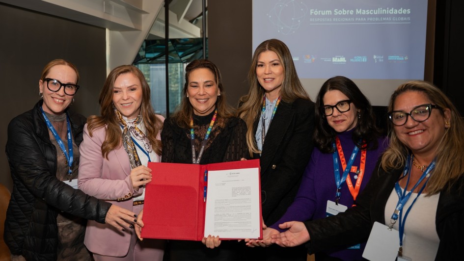 A fotografia registra um grupo de seis mulheres durante um evento internacional na ONU, celebrando a assinatura de um termo de coopera&ccedil;&atilde;o entre os estados do COSUD (Cons&oacute;rcio de Integra&ccedil;&atilde;o Sul e Sudeste) para o fortalecimento de pol&iacute;ticas para mulheres.

Elementos Principais
O Grupo: Seis mulheres est&atilde;o posicionadas lado a lado, em plano m&eacute;dio, sorrindo para a c&acirc;mera. Elas possuem diferentes perfis e estilos, vestindo blazers, jaquetas e roupas sociais em tons de preto, rosa e roxo. Todas utilizam crach&aacute;s com cord&otilde;es coloridos t&iacute;picos de confer&ecirc;ncias internacionais.

O Documento: No centro, tr&ecirc;s das mulheres seguram juntas uma pasta de couro vermelha aberta, exibindo um documento oficial de uma p&aacute;gina com o bras&atilde;o do Estado do Rio Grande do Sul no topo.

A Proje&ccedil;&atilde;o: Ao fundo, &agrave; direita, uma tela exibe o t&iacute;tulo do evento: "F&oacute;rum Sobre Masculinidades - RESPOSTAS REGIONAIS PARA PROBLEMAS GLOBAIS". Abaixo do t&iacute;tulo, h&aacute; logotipos institucionais, incluindo o da ONU Mulheres e do Governo do Brasil.

Detalhes e Ambiente
Cen&aacute;rio: O ambiente &eacute; sofisticado e moderno. &Agrave; esquerda, destaca-se uma estrutura arquitet&ocirc;nica com janelas de vidro que mostram o reflexo de pr&eacute;dios urbanos, sugerindo a sede da ONU em Nova York.

Composi&ccedil;&atilde;o: A imagem foca na uni&atilde;o das m&atilde;os segurando o documento vermelho, simbolizando a coopera&ccedil;&atilde;o e o compromisso firmado entre as gestoras dos diferentes estados.

Ilumina&ccedil;&atilde;o: A luz &eacute; suave e bem distribu&iacute;da, ressaltando os detalhes dos rostos e a formalidade do encontro.