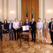 A foto mostra um grupo alinhado para registro oficial em um sal&atilde;o hist&oacute;rico, com o governador ao centro, que segura um documento aberto sobre uma mesa. Ao fundo, duas bandeiras est&atilde;o posicionadas diante da cortina dourada.