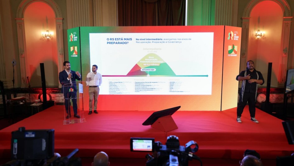 A foto mostra o governador Eduardo Leite e o vice-governador Gabriel Souza em um palco, com um tel&atilde;o ao fundo com informa&ccedil;&otilde;es sobre o Plano Rio Grande.
