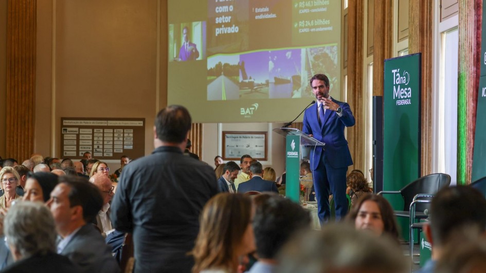 A imagem mostra um evento realizado em um sal&atilde;o hist&oacute;rico amplo. O p&uacute;blico est&aacute; sentado em mesas redondas, enquanto o governador se dirige aos participantes a partir de um p&uacute;lpito transparente. Ao fundo, um tel&atilde;o exibe apresenta&ccedil;&atilde;o com o tema &ldquo;Parcerias com o setor privado&rdquo;, acompanhada de imagens e textos institucionais. Pain&eacute;is verdes com a marca T&aacute; na Mesa &ndash; Federasul comp&otilde;em a cenografia.