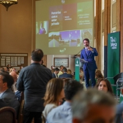 A imagem mostra um evento realizado em um sal&atilde;o hist&oacute;rico amplo. O p&uacute;blico est&aacute; sentado em mesas redondas, enquanto o governador se dirige aos participantes a partir de um p&uacute;lpito transparente. Ao fundo, um tel&atilde;o exibe apresenta&ccedil;&atilde;o com o tema &ldquo;Parcerias com o setor privado&rdquo;, acompanhada de imagens e textos institucionais. Pain&eacute;is verdes com a marca T&aacute; na Mesa &ndash; Federasul comp&otilde;em a cenografia.