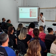A imagem mostra uma sala cheia de pessoas sentadas assistindo a uma palestra. Um homem est&aacute; em p&eacute;, falando com o p&uacute;blico. Ao fundo, h&aacute; um projeto de slides com a apresenta&ccedil;&atilde;o, uma mesa e um quadro branco.
