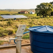 A imagem mostra uma caixa d'&aacute;gua azul em primeiro plano e, ao fundo, uma paisagem rural, com campos, uma casa e uma capela. Tamb&eacute;m h&aacute; placas solares e &aacute;rvores em um dia ensolarado. 