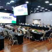 A imagem mostra um grande audit&oacute;rio preparado para um encontro institucional, com mesas retangulares dispostas em formato de &ldquo;U&rdquo; e ocupadas por participantes que utilizam notebooks, pastas e garrafas de &aacute;gua. No fundo do sal&atilde;o, um tel&atilde;o amplo exibe a identidade visual do evento: &ldquo;ENCAT Mato Grosso&rdquo;, com cores verde, azul e amarelo.
