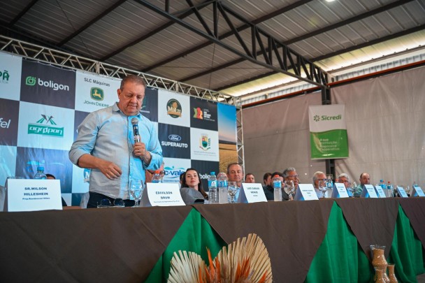 Um homem de camisa azul claro fala ao microfone em pé, numa mesa de autoridades em evento agropecuário coberto. Ao fundo, banner com patrocinadores. Sua plaquinha identifica: Edivilson Brum, Secretário de Agricultura.