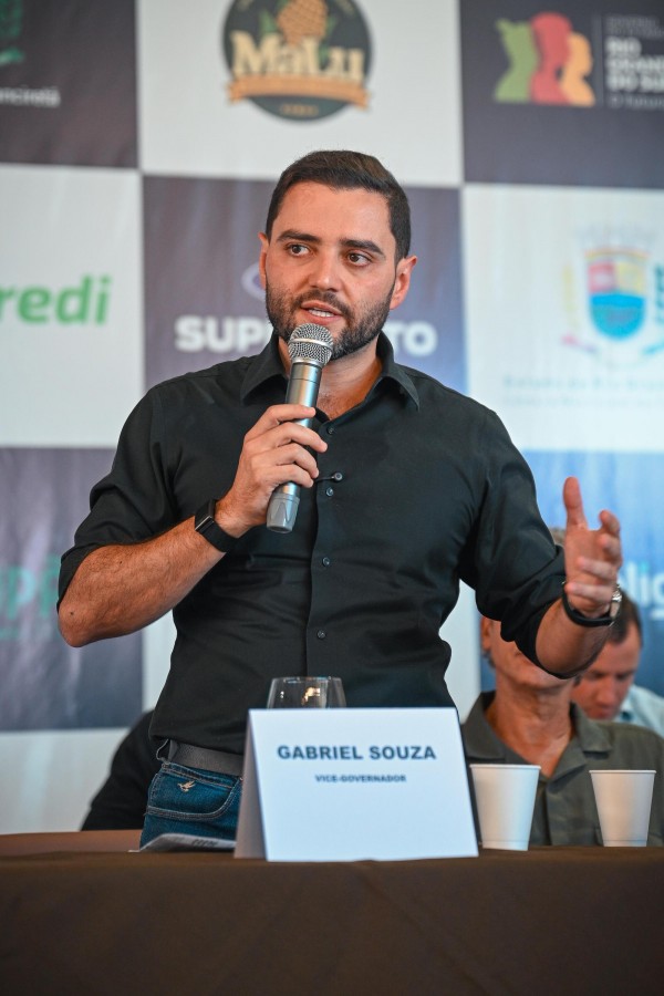 Homem jovem de camisa preta discursa ao microfone com expressão animada e gesto com a mão. À sua frente, plaquinha com "Gabriel Souza – Vice-Governador". Ao fundo, logos de empresas.