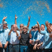 Grupo de cerca de dez pessoas joga gr&atilde;os de soja para o alto em comemora&ccedil;&atilde;o, sob c&eacute;u azul intenso. V&aacute;rios usam chap&eacute;us de palha e camisas claras. No centro, destaca-se um homem de preto. A cena celebra a abertura da colheita da soja.