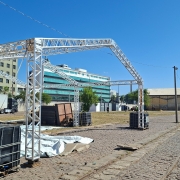 A fotografia registra os preparativos estruturais no Cais Mau&aacute;, em Porto Alegre, para a realiza&ccedil;&atilde;o do evento de inova&ccedil;&atilde;o South Summit Brazil 2026.

Elementos Principais
Estrutura Met&aacute;lica: Em primeiro plano, destaca-se a arma&ccedil;&atilde;o de uma grande tenda ou pavilh&atilde;o feita de treli&ccedil;as de alum&iacute;nio (box truss). A estrutura est&aacute; em fase de montagem, posicionada sobre o cal&ccedil;amento de paralelep&iacute;pedos.

Base de Sustenta&ccedil;&atilde;o: Os pilares da estrutura est&atilde;o fixados e estabilizados por grandes reservat&oacute;rios pl&aacute;sticos pretos (IBCs) cheios de &aacute;gua, que servem como contrapesos.

Trabalhador: &Agrave; esquerda, um oper&aacute;rio de costas, vestindo camiseta preta e capacete de seguran&ccedil;a branco, observa a montagem.

Cen&aacute;rio e Ambiente
Cais Mau&aacute;: &Agrave; direita, estendem-se os trilhos hist&oacute;ricos de trem e a linha de armaz&eacute;ns amarelos caracter&iacute;sticos do porto, que margeiam o Lago Gua&iacute;ba (vis&iacute;vel ao fundo, &agrave; extrema direita).

Entorno Urbano: Ao fundo, o cen&aacute;rio &eacute; composto por pr&eacute;dios comerciais, incluindo um edif&iacute;cio com fachada de vidro verde e outros de arquitetura mais tradicional. Um caminh&atilde;o branco est&aacute; estacionado na &aacute;rea de log&iacute;stica &agrave; esquerda.

Condi&ccedil;&otilde;es Clim&aacute;ticas: O dia est&aacute; ensolarado, com c&eacute;u azul sem nuvens, o que proporciona uma ilumina&ccedil;&atilde;o clara e sombras n&iacute;tidas no ch&atilde;o.

Composi&ccedil;&atilde;o e Contexto
A imagem utiliza uma perspectiva que valoriza a extens&atilde;o do Cais Mau&aacute;, conectando a infraestrutura tempor&aacute;ria do evento ao patrim&ocirc;nio hist&oacute;rico da cidade.

O registro enfatiza a fase de "ajustes finais" e montagem log&iacute;stica para receber o p&uacute;blico do festival de inova&ccedil;&atilde;o.