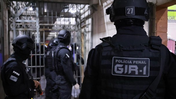 Foto mostra policiais penais, de costa, e com uniformes pretos e capacetes, no corredor do Complexo Prisional de Canoas, ao fundo aparecem grades no meio do corredor. 