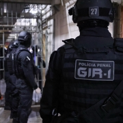 Foto mostra policiais penais, de costa, e com uniformes pretos e capacetes, no corredor do Complexo Prisional de Canoas, ao fundo aparecem grades no meio do corredor. 