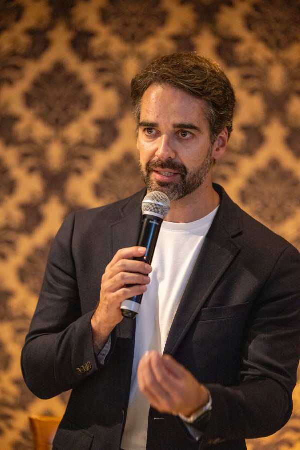 A fotografia é um plano médio e fechado que captura o governador Eduardo Leite em um momento de discurso ou explicação durante um evento institucional.

Elementos Principais
O Governador: Eduardo Leite, com barba aparada e cabelo curto, está posicionado no centro da imagem. Ele veste um traje esporte fino, composto por um blazer azul-marinho de botões sobre uma camiseta branca básica.

A Ação: Ele segura um microfone com a mão direita e gesticula com a mão esquerda à frente do corpo, com os dedos semi-abertos, como se estivesse enfatizando um ponto ou explicando algo ao público. Sua expressão facial é focada e serena, com o olhar direcionado para a esquerda da câmera.

O Ambiente: O fundo é composto por um painel de parede com uma textura de tecido dourado e um padrão de damasco clássico e elegante. A iluminação é suave e uniforme, típica de um evento corporativo ou conferência.

Contexto
A imagem registra a participação do governador na cerimônia de lançamento da pedra fundamental do Club Med Gramado, reforçando o compromisso do Estado com o turismo e o desenvolvimento econômico.