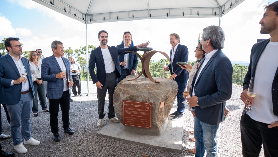A fotografia registra o ato solene de lan&ccedil;amento da pedra fundamental do resort Club Med Gramado, com a participa&ccedil;&atilde;o do governador Eduardo Leite e executivos do empreendimento.

Elementos Principais
O Monumento: No centro da imagem, sobre uma base de concreto, repousa uma grande rocha natural (pedra fundamental). Nela, est&aacute; fixada uma placa met&aacute;lica avermelhada com inscri&ccedil;&otilde;es comemorativas. Sobre a pedra, h&aacute; uma escultura de metal composta por dois arcos que se cruzam.

O Governador: Eduardo Leite, vestindo um blazer azul-marinho sobre uma camiseta branca e cal&ccedil;a cinza, est&aacute; posicionado atr&aacute;s da pedra. Ele segura um pequeno objeto cil&iacute;ndrico (provavelmente uma c&aacute;psula do tempo) que est&aacute; sendo depositado no topo do monumento.

As Autoridades e Convidados: Cerca de oito homens e uma mulher cercam o monumento sob uma tenda. Eles vestem trajes esporte fino (blazers e camisas sociais). Alguns seguram ta&ccedil;as com espumante, celebrando o momento com sorrisos.

Cen&aacute;rio e Ambiente
Local: O evento ocorre em uma &aacute;rea elevada e aberta. Ao fundo, avista-se uma vasta paisagem natural caracter&iacute;stica da Serra Ga&uacute;cha, com muitas copas de &aacute;rvores (arauc&aacute;rias) e o horizonte sob um c&eacute;u azul com nuvens esparsas.

Estrutura: O ato &eacute; protegido por uma tenda de lona branca. O ch&atilde;o &eacute; coberto por brita cinza.

Composi&ccedil;&atilde;o: A foto captura o grupo por inteiro, destacando a integra&ccedil;&atilde;o entre o investimento privado e o cen&aacute;rio natural da regi&atilde;o.