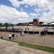A fotografia registra a estrutura da maior pista de skate da Am&eacute;rica Latina, na Orla do Gua&iacute;ba, preparada para a abertura do STU National 2026 em Porto Alegre.

Elementos Principais
A Pista (Skatepark): Em plano m&eacute;dio, v&ecirc;-se a vasta &aacute;rea de concreto liso composta por diferentes obst&aacute;culos de modalidades como Street e Park, incluindo rampas, corrim&atilde;os e v&atilde;os t&eacute;cnicos. O asfalto exibe a inscri&ccedil;&atilde;o "STU NATIONAL PORTO ALEGRE" centralizada no piso.

Estrutura de Evento: Ao fundo, destacam-se grandes tendas brancas, arquibancadas met&aacute;licas laterais e uma torre central com um tel&atilde;o de LED e o logotipo do evento em fundo vermelho. Diversos ombrelones (guarda-s&oacute;is) azuis da marca Red Bull est&atilde;o espalhados pela &aacute;rea t&eacute;cnica.

Pessoas e Movimenta&ccedil;&atilde;o: Algumas poucas pessoas, entre skatistas e equipe t&eacute;cnica, circulam pela pista. Em primeiro plano, dois jovens de camisetas brancas e bon&eacute;s observam a pista apoiados em uma grade met&aacute;lica de conten&ccedil;&atilde;o.

Detalhes e Ambiente
Cen&aacute;rio: A imagem &eacute; capturada a partir de um ponto elevado (provavelmente o topo da arquibancada de concreto). O horizonte &eacute; marcado por &aacute;rvores verdes e o c&eacute;u est&aacute; azul, densamente povoado por nuvens brancas e volumosas.

Ilumina&ccedil;&atilde;o: A luz solar &eacute; difusa devido &agrave;s nuvens, criando uma ilumina&ccedil;&atilde;o clara e uniforme sobre todo o complexo esportivo.