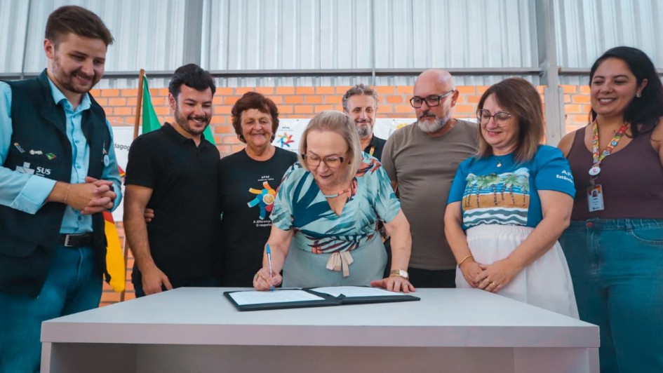 A fotografia registra o momento da assinatura oficial para a habilita&ccedil;&atilde;o de um Centro de Atendimento em Sa&uacute;de (CAS) do programa TEAcolhe em Est&acirc;ncia Velha, voltado ao atendimento especializado para pessoas com autismo.

Elementos Principais
O Ato da Assinatura: No centro da imagem, a Secret&aacute;ria da Sa&uacute;de do Estado, Arita Bergmann (vestindo uma blusa estampada em tons de azul e verde e cal&ccedil;a clara), est&aacute; inclinada sobre uma mesa cinza, assinando um documento em uma pasta preta.

Autoridades e Gestores: Sete pessoas cercam a secret&aacute;ria, acompanhando o ato com express&otilde;es de satisfa&ccedil;&atilde;o e sorrisos.

&Agrave; esquerda, o homem de colete preto do SUS/Secretaria da Sa&uacute;de observa com as m&atilde;os entrela&ccedil;adas.

Ao lado dele, um homem de camiseta polo preta e uma mulher com camiseta da APAE (onde se l&ecirc; "A diferen&ccedil;a, o respeito") acompanham a assinatura.

&Agrave; direita, um homem de barba grisalha e camiseta marrom, uma mulher com camiseta azul estampada e uma jovem de regata bord&ocirc; completam o grupo.

Cen&aacute;rio e Ambiente
Local: O evento ocorre em um ambiente interno de aspecto industrial ou em obras, com paredes de tijolos aparentes e teto alto de estrutura met&aacute;lica galvanizada.

Identifica&ccedil;&atilde;o: Ao fundo, &agrave; esquerda, &eacute; vis&iacute;vel parte de uma bandeira do Rio Grande do Sul.

Ilumina&ccedil;&atilde;o: A luz &eacute; frontal e clara, destacando o documento sobre a mesa e as fei&ccedil;&otilde;es dos participantes, refor&ccedil;ando o car&aacute;ter comunit&aacute;rio e institucional da entrega.