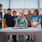 A fotografia registra o momento da assinatura oficial para a habilita&ccedil;&atilde;o de um Centro de Atendimento em Sa&uacute;de (CAS) do programa TEAcolhe em Est&acirc;ncia Velha, voltado ao atendimento especializado para pessoas com autismo.

Elementos Principais
O Ato da Assinatura: No centro da imagem, a Secret&aacute;ria da Sa&uacute;de do Estado, Arita Bergmann (vestindo uma blusa estampada em tons de azul e verde e cal&ccedil;a clara), est&aacute; inclinada sobre uma mesa cinza, assinando um documento em uma pasta preta.

Autoridades e Gestores: Sete pessoas cercam a secret&aacute;ria, acompanhando o ato com express&otilde;es de satisfa&ccedil;&atilde;o e sorrisos.

&Agrave; esquerda, o homem de colete preto do SUS/Secretaria da Sa&uacute;de observa com as m&atilde;os entrela&ccedil;adas.

Ao lado dele, um homem de camiseta polo preta e uma mulher com camiseta da APAE (onde se l&ecirc; "A diferen&ccedil;a, o respeito") acompanham a assinatura.

&Agrave; direita, um homem de barba grisalha e camiseta marrom, uma mulher com camiseta azul estampada e uma jovem de regata bord&ocirc; completam o grupo.

Cen&aacute;rio e Ambiente
Local: O evento ocorre em um ambiente interno de aspecto industrial ou em obras, com paredes de tijolos aparentes e teto alto de estrutura met&aacute;lica galvanizada.

Identifica&ccedil;&atilde;o: Ao fundo, &agrave; esquerda, &eacute; vis&iacute;vel parte de uma bandeira do Rio Grande do Sul.

Ilumina&ccedil;&atilde;o: A luz &eacute; frontal e clara, destacando o documento sobre a mesa e as fei&ccedil;&otilde;es dos participantes, refor&ccedil;ando o car&aacute;ter comunit&aacute;rio e institucional da entrega.