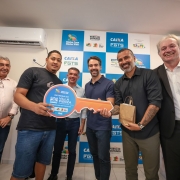 A imagem mostra um grupo reunido em um ambiente interno durante a entrega simb&oacute;lica de moradias. No centro, v&aacute;rias pessoas seguram uma chave cenogr&aacute;fica gigante, identificada com o texto &ldquo;Entrega do Residencial Villa Pienza &ndash; PAC / Minha Casa Minha Vida&rdquo;. O fundo &eacute; composto por um painel com logomarcas da Caixa, FGTS, Minha Casa Minha Vida e Governo Federal.