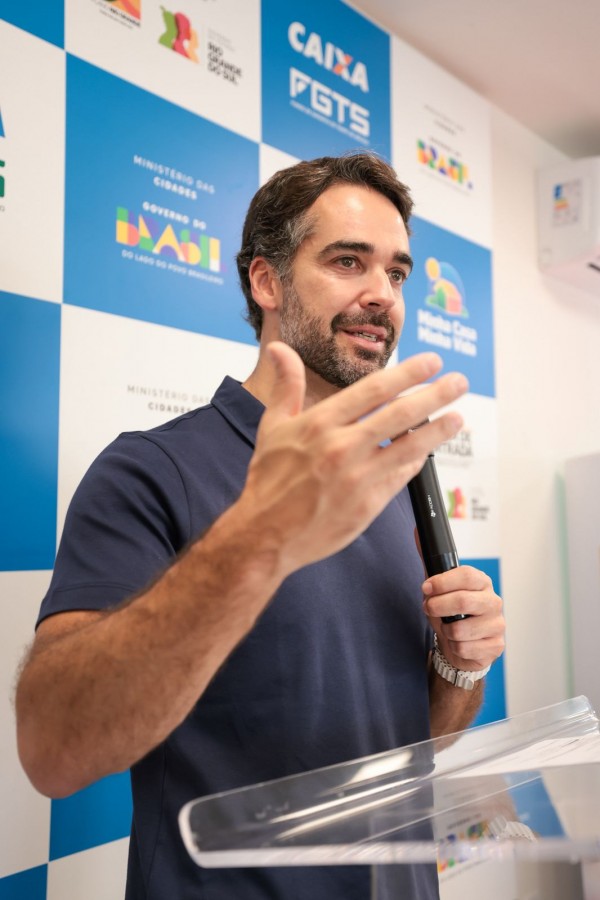 A foto mostra o governador fazendo discurso em um púlpito transparente diante de um painel institucional com logomarcas da Caixa, FGTS, Minha Casa Minha Vida, e Governo Federal.