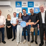 A imagem mostra um grupo reunido em um ambiente interno durante a entrega simb&oacute;lica de moradias. No centro, v&aacute;rias pessoas seguram uma chave cenogr&aacute;fica gigante, identificada com o texto &ldquo;Entrega do Residencial Villa Pienza &ndash; PAC / Minha Casa Minha Vida&rdquo;. O fundo &eacute; composto por um painel com logomarcas da Caixa, FGTS, Minha Casa Minha Vida e Governo Federal.