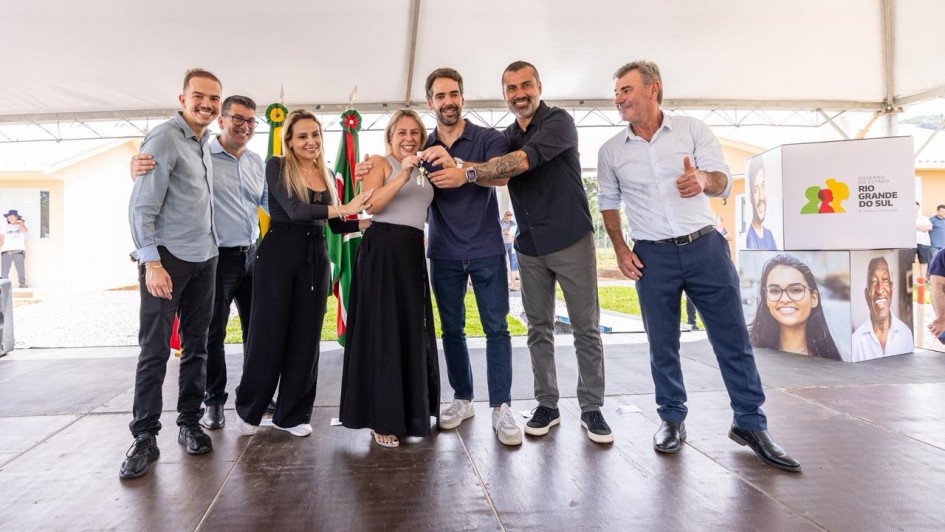 A fotografia registra o governador Eduardo Leite durante a entrega de 28 casas definitivas em Salvador do Sul, constru&iacute;das por meio do programa "A Casa &eacute; Sua - Calamidade".

Elementos Principais
O Governador: Eduardo Leite est&aacute; em primeiro plano, &agrave; esquerda. Ele veste uma camisa social azul clara com as mangas dobradas e cal&ccedil;a cinza. Ele sorri enquanto gesticula com as m&atilde;os, interagindo com os moradores e autoridades presentes.

A Entrega Simb&oacute;lica: No centro da imagem, o governador segura, junto com uma mulher (benefici&aacute;ria do programa), uma chave simb&oacute;lica de grandes dimens&otilde;es. A chave &eacute; feita de material r&iacute;gido branco e azul, com o logotipo do Governo do Estado do Rio Grande do Sul e a inscri&ccedil;&atilde;o "A Casa &eacute; Sua".

Os Participantes: Ao redor deles, um grupo de pessoas acompanha o ato com alegria. &Agrave; direita, um homem de camisa polo azul e uma mulher de blusa branca sorriem e aplaudem o momento da entrega.

Cen&aacute;rio e Ambiente
As Moradias: Ao fundo, s&atilde;o vis&iacute;veis as novas casas do conjunto habitacional. As unidades s&atilde;o de alvenaria, pintadas em tons de terracota e bege, com telhados de fibrocimento cinza e janelas brancas.

Infraestrutura: O evento ocorre na rua em frente &agrave;s casas, que possui pavimenta&ccedil;&atilde;o de blocos de concreto (intertravados). O dia est&aacute; ensolarado, com c&eacute;u azul e poucas nuvens, destacando as cores vibrantes das novas moradias.

Simbolismo: A cena captura um momento de dignidade e recome&ccedil;o para as fam&iacute;lias beneficiadas pelo programa habitacional ap&oacute;s eventos de calamidade.