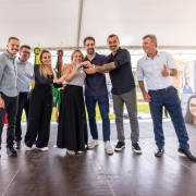 A fotografia registra o governador Eduardo Leite durante a entrega de 28 casas definitivas em Salvador do Sul, constru&iacute;das por meio do programa "A Casa &eacute; Sua - Calamidade".

Elementos Principais
O Governador: Eduardo Leite est&aacute; em primeiro plano, &agrave; esquerda. Ele veste uma camisa social azul clara com as mangas dobradas e cal&ccedil;a cinza. Ele sorri enquanto gesticula com as m&atilde;os, interagindo com os moradores e autoridades presentes.

A Entrega Simb&oacute;lica: No centro da imagem, o governador segura, junto com uma mulher (benefici&aacute;ria do programa), uma chave simb&oacute;lica de grandes dimens&otilde;es. A chave &eacute; feita de material r&iacute;gido branco e azul, com o logotipo do Governo do Estado do Rio Grande do Sul e a inscri&ccedil;&atilde;o "A Casa &eacute; Sua".

Os Participantes: Ao redor deles, um grupo de pessoas acompanha o ato com alegria. &Agrave; direita, um homem de camisa polo azul e uma mulher de blusa branca sorriem e aplaudem o momento da entrega.

Cen&aacute;rio e Ambiente
As Moradias: Ao fundo, s&atilde;o vis&iacute;veis as novas casas do conjunto habitacional. As unidades s&atilde;o de alvenaria, pintadas em tons de terracota e bege, com telhados de fibrocimento cinza e janelas brancas.

Infraestrutura: O evento ocorre na rua em frente &agrave;s casas, que possui pavimenta&ccedil;&atilde;o de blocos de concreto (intertravados). O dia est&aacute; ensolarado, com c&eacute;u azul e poucas nuvens, destacando as cores vibrantes das novas moradias.

Simbolismo: A cena captura um momento de dignidade e recome&ccedil;o para as fam&iacute;lias beneficiadas pelo programa habitacional ap&oacute;s eventos de calamidade.