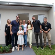 Grupo posicionado em frente a uma casa t&eacute;rrea rec&eacute;m-constru&iacute;da, com fachada clara, porta branca e janela lateral. As pessoas, incluindo o governador, est&atilde;o alinhadas para foto sobre uma &aacute;rea gramada e cal&ccedil;ada de acesso.