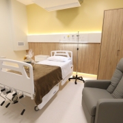 Imagem de um quarto hospitalar com cama el&eacute;trica, poltrona reclin&aacute;vel, suporte para soro, arm&aacute;rio e pia. O ambiente &eacute; moderno, bem iluminado e organizado, com detalhes em madeira e luz indireta na parede.