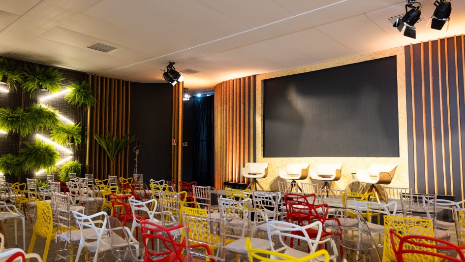 O ambiente mostra um audit&oacute;rio preparado para palestras, com cadeiras coloridas distribu&iacute;das em frente a um palco. O palco possui uma tela grande ao centro, quatro cadeiras altas e decora&ccedil;&atilde;o com pain&eacute;is de madeira vertical.